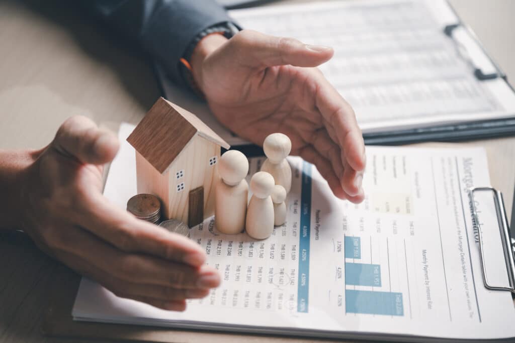 Family insurance plan protect. Help medicine protection. Father mother children insurance. A person is holding a small wooden house and three figurines on top of a piece of paper.